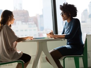Zwei Studentinnen sitzen sich an einem Tisch vor einem Fenster gegenüber und sind in ein Gespräch vertieft.