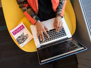 Eine Person von oben, die an einem Laptop schreibt. Danaben liegt auf dem Tisch ein Python Buch.