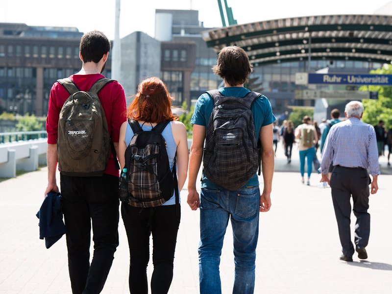 Drei Personen von hinten, die über die Uni-Brücke in Richtung Campus gehen.