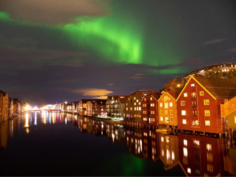 Häuserfronten an einem Fluss in der Dunkelheit, darüber Polarlichter am Himmel.